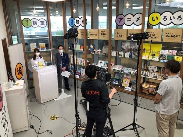 J:COMチャンネル生番組「LIVEニュース」のアナウンサー・仁科美咲さん。角川武蔵野ミュージアムを背景に笑顔で両手を振っている。