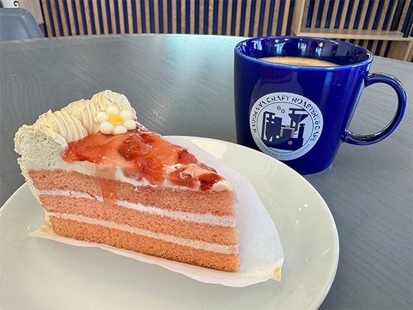 目にも鮮やかなピンク色の生地と生クリームが層になったケーキの画像。奥にはホットの飲み物が入った青い陶器のカップがある。